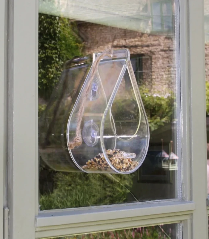 A clear Perspex Wildlife World Dewdrop window bird feeder with a feeding tray, attached to a window pane with rubber suction cups, and a perch for birds to land on.